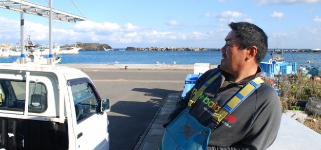 笑顔で話してくれる小松さんのバックには、津波で壊されたままの消波ブロックが見える